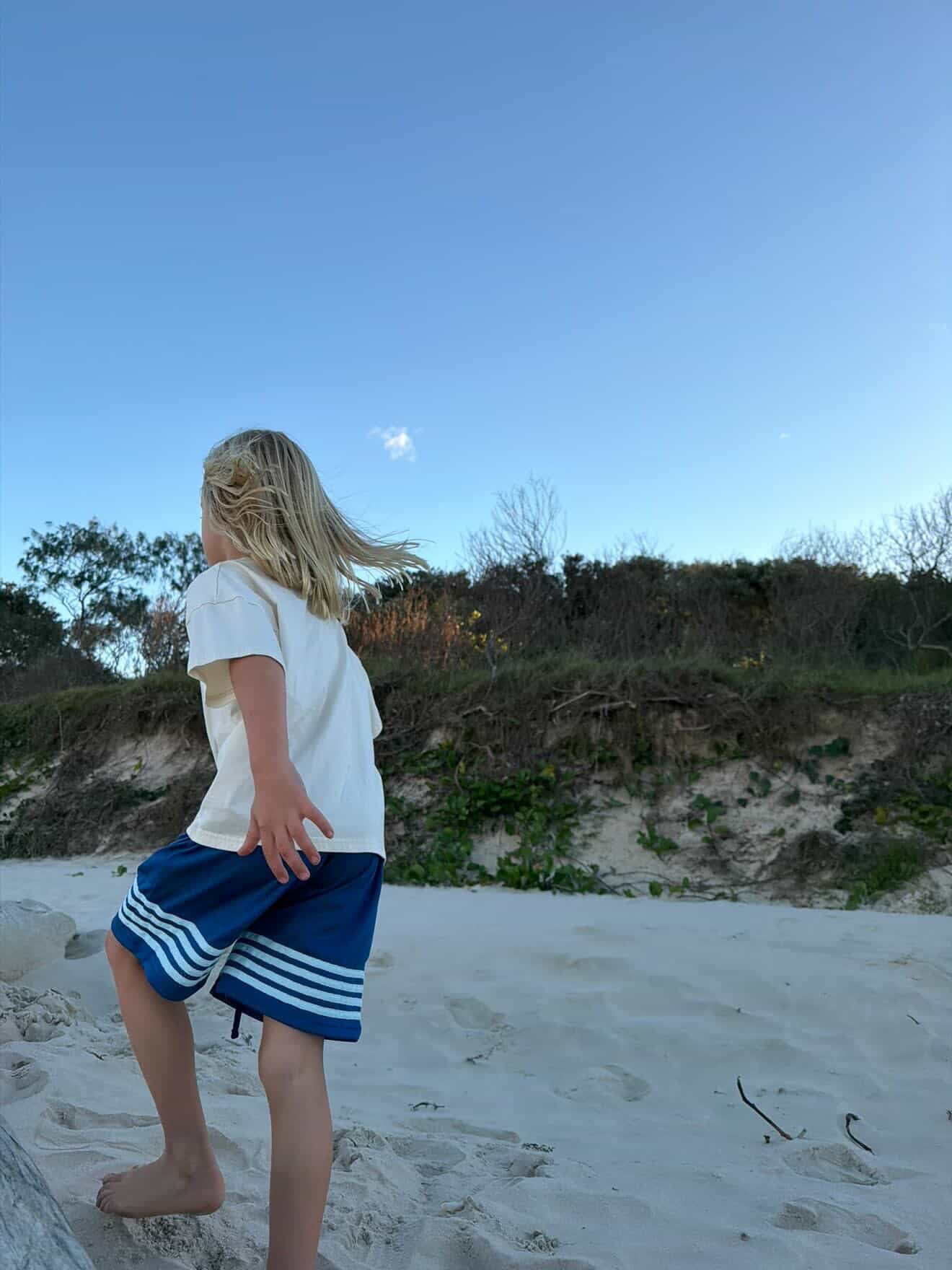 A young child with blonde hair seen from behind, standing on a sandy beach wearing a white t-shirt and blue athletic shorts with white stripes, looking out towards a dune and blue sky.