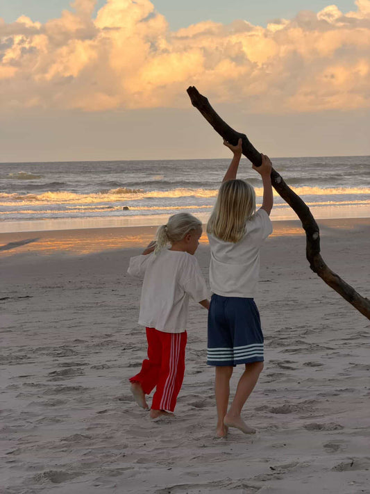 Two children walking on a sandy beach at sunset, with one holding a large piece of driftwood overhead. The child on the right wears a white t-shirt and navy shorts with white stripes, while the child on the left wears a white top and red pants with white side stripes.